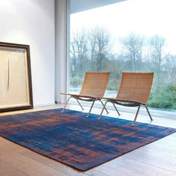 Louis De Poortere Venetian Dust rug in blue and rust tones placed in modern room with wooden flooring and wicker chairs