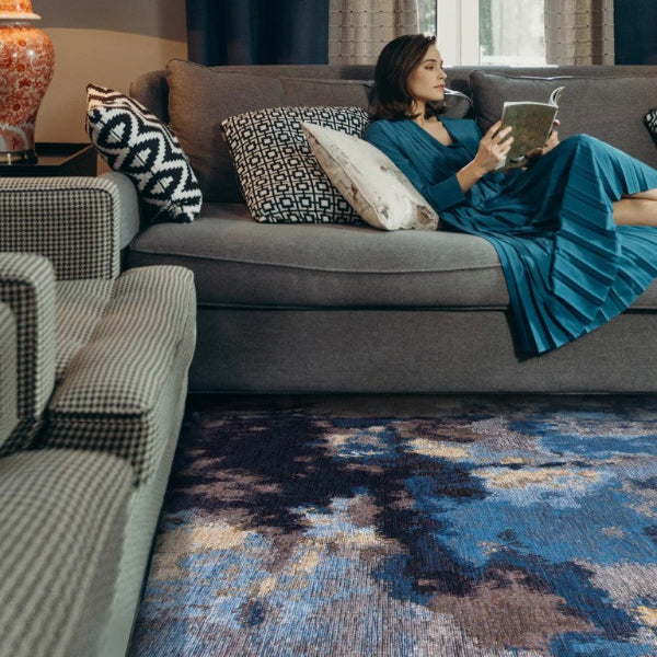 Woman in a blue dress sitting on a gray sofa reading a book in a living room.
